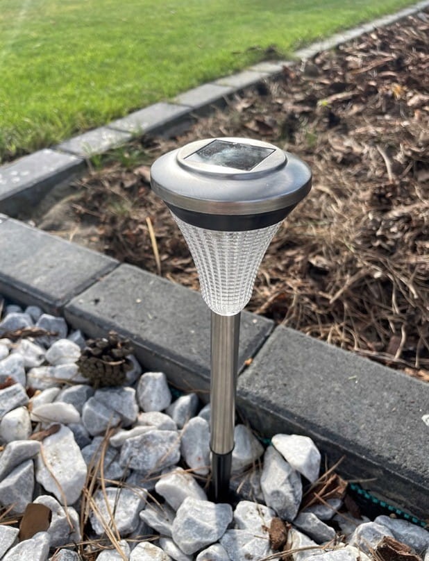Lampa solarna ogrodowa Latarnia III, wbijana, wysokość 32 cm, z metalowym srebrnym wykończeniem i przezroczystą, teksturowaną osłoną. Zainstalowana wśród białych kamieni na trawniku, z panelem słonecznym na górze.