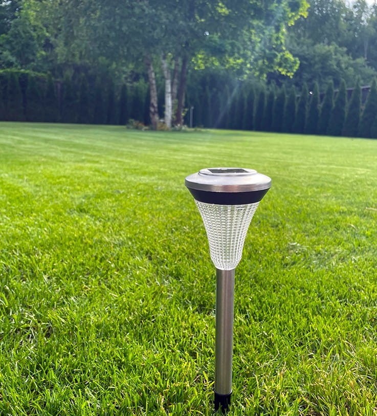 Srebrna lampa solarna ogrodowa, wbijana, o wysokości 32 cm. Ujęcie z boku, na zielonym trawniku. Podstawa metalowa, klosz z teksturowanego tworzywa. Idealna do oświetlenia ogrodu lub ścieżek.