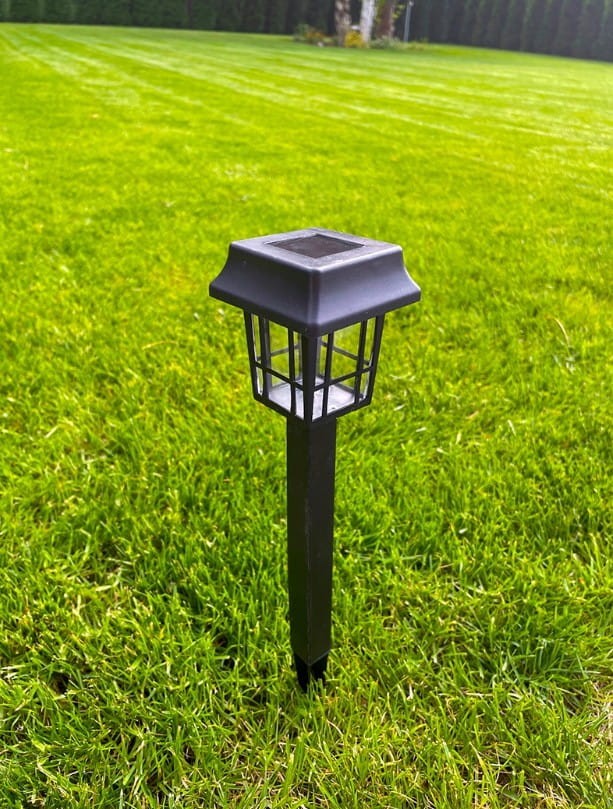 Latarnia IV, lampa solarna ogrodowa w kolorze czarnym, wbijana, o wysokości 32 cm. Posiada kwadratowy daszek z przezroczystymi ściankami, umieszczona na prostym, metalowym słupku, stojąca na zielonym trawniku.