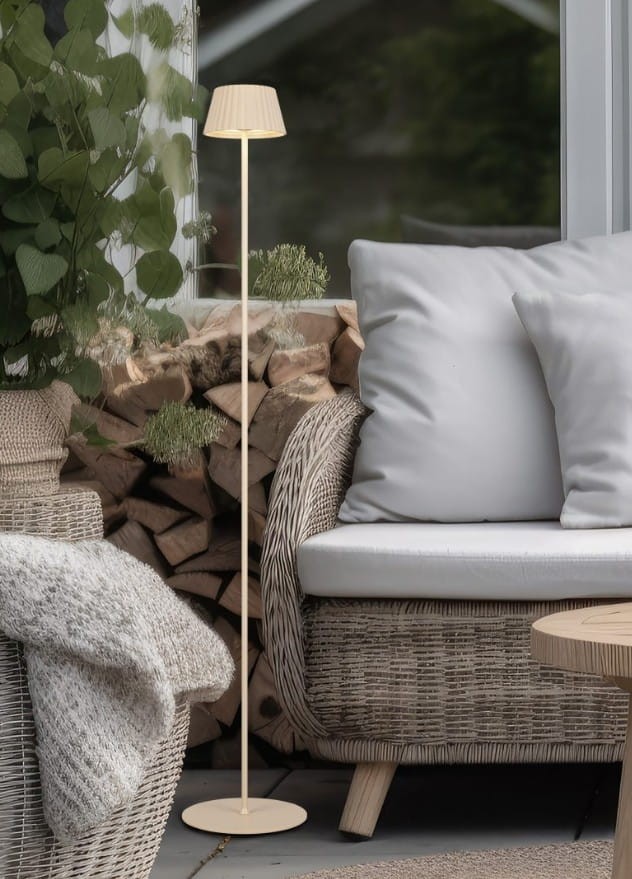 Lampa stojąca zewnętrzna Suarez w kolorze piaskowym, z abażurem o fakturze. Umieszczona obok sofy na tarasie, z subtelnym oświetleniem LED 1,5W. W tle widoczne drewno i rośliny, tworzące przytulną atmosferę.