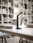 Lampa stołowa Skip bezprzewodowa, czarna, z wbudowanym źródłem LED 2W. Ustawiona na białym biurku, z prostokątnym kloszem i minimalistycznym designem. W tle regał z książkami.
