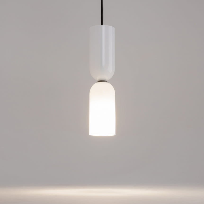 Wisząca lampa Memory z jednym gniazdem E14, w kolorze białym. Składa się z gładkiego, błyszczącego abażura oraz matowego klosza, emitującego ciepłe światło. Produkt zawieszony na czarnym przewodzie, na jednolitym tle.