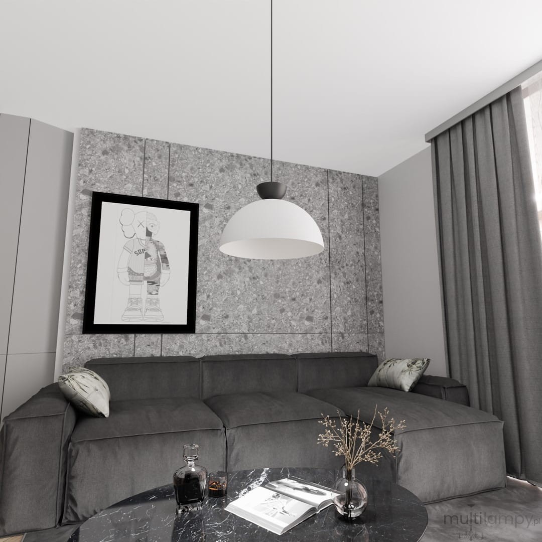 Lampa wisząca Lunaro L w kolorze białym z czarnym detalem, zawieszona nad szarym narożnikiem w nowoczesnym wnętrzu. Tło stanowi szara ściana z teksturą oraz obraz w czarnej ramie. Na stole widoczne książki i dekoracja w wazonie.