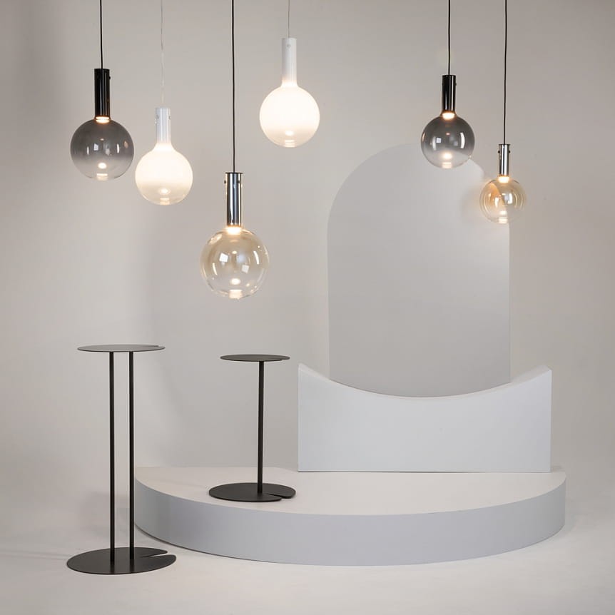 Lampa wisząca Nebula D25, czarna, z jedną żarówką LED 6W. Zawieszona wśród innych lamp w różnych kolorach i kształtach, na tle minimalistycznej aranżacji z białymi elementami oraz dwoma stojącymi stolikami o czarnych podstawach.