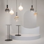 Lampa wisząca Nebula D25, czarna, z jedną żarówką LED 6W. Zawieszona wśród innych lamp w różnych kolorach i kształtach, na tle minimalistycznej aranżacji z białymi elementami oraz dwoma stojącymi stolikami o czarnych podstawach.