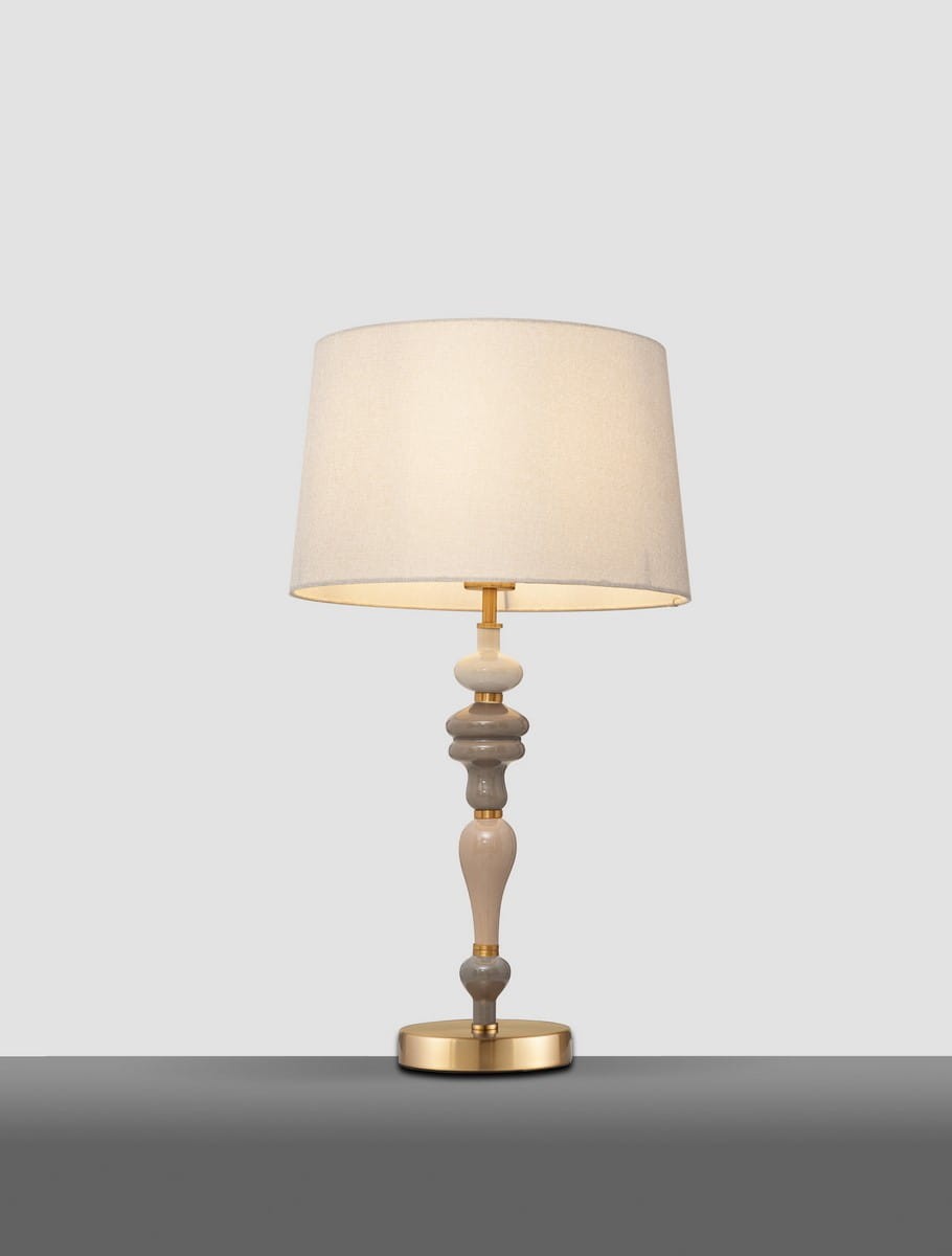 Lampa stołowa Alem w kolorze antycznego mosiądzu i beżu, z cylindrycznym abażurem. Podstawa z dekoracyjnymi elementami w różnych odcieniach beżu i szarości. Ujęcie frontalne, produkt stojący na szarej powierzchni.