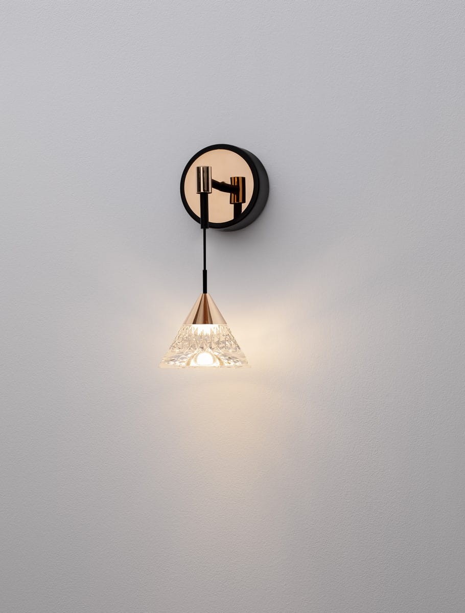 Kinkiet Guay 1xLED/7W w kolorze czarnym i złotym. Okrągła, czarna podstawa z metalowym mocowaniem, zwisająca, przezroczysta klosz w kształcie stożka, ozdobnie teksturowany, emitujący ciepłe światło. Zainstalowany na jasnej ścianie.