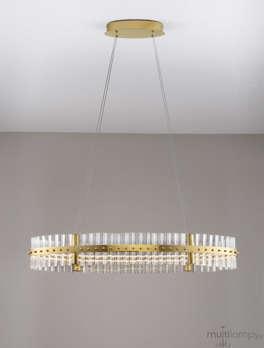 Lampa wisząca Mayo D90 z jedną żarówką LED 45W w złotej oprawie. Posiada półokrągły kształt, z przezroczystymi elementami szklanymi ułożonymi w rzędy. Zawieszona na dwóch cienkich linkach, montowana do sufitu. Tło w jasnoszarym kolorze.