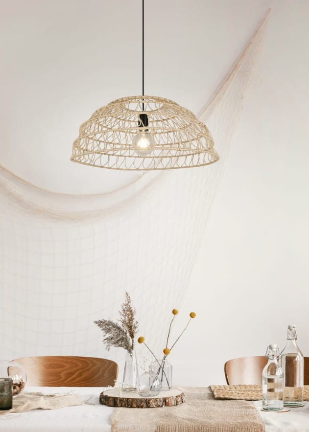 Lampa wisząca Paulette z materiału w kolorze jasnobrązowym, z czarnym zawieszeniem, o okrągłym kształcie. Widoczna żarówka E27. Produkt zawieszony nad stołem, w minimalistycznym otoczeniu z naturalnymi dekoracjami.