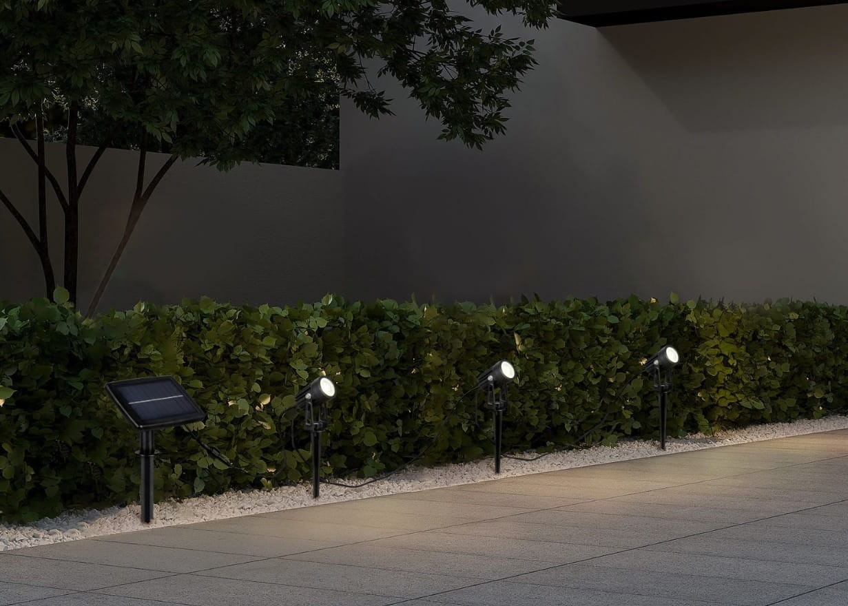 Zestaw trzech solarnych opraw dogruntowych Obidos Hybrid, czarne, z trzema diodami LED 1,5W, 4000K. Ujęcie z boku, oprawy ustawione wzdłuż żywopłotu na jasnym podłożu. Widoczny panel solarny obok jednej z opraw.