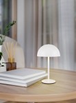 Lampa stołowa Munoz w kolorze białym, z okrągłym kloszem i cienką nóżką, umieszczona na drewnianym stole obok stosu białych książek. W tle widoczne rośliny i zasłony, co tworzy przytulną aranżację wnętrza.
