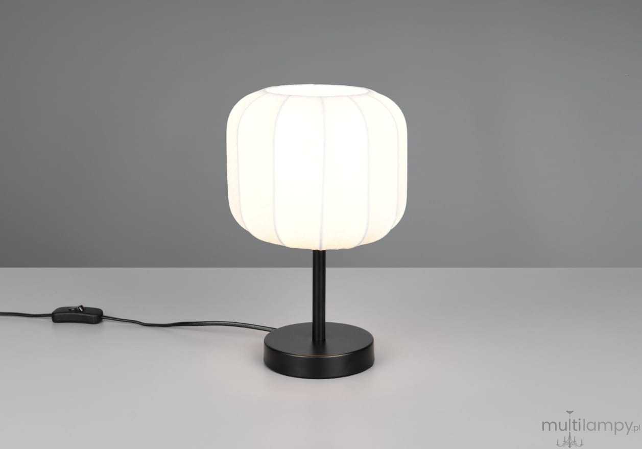 Lampa stołowa Madeline z czarną podstawą i białym kloszem o cylindrycznym kształcie, stojąca na płaskiej powierzchni. Klosz wykonany z materiału z widocznymi przeszyciami, z kablem z włącznikiem. Tło szare, proste.