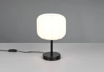 Lampa stołowa Madeline z czarną podstawą i białym kloszem o cylindrycznym kształcie, stojąca na płaskiej powierzchni. Klosz wykonany z materiału z widocznymi przeszyciami, z kablem z włącznikiem. Tło szare, proste.