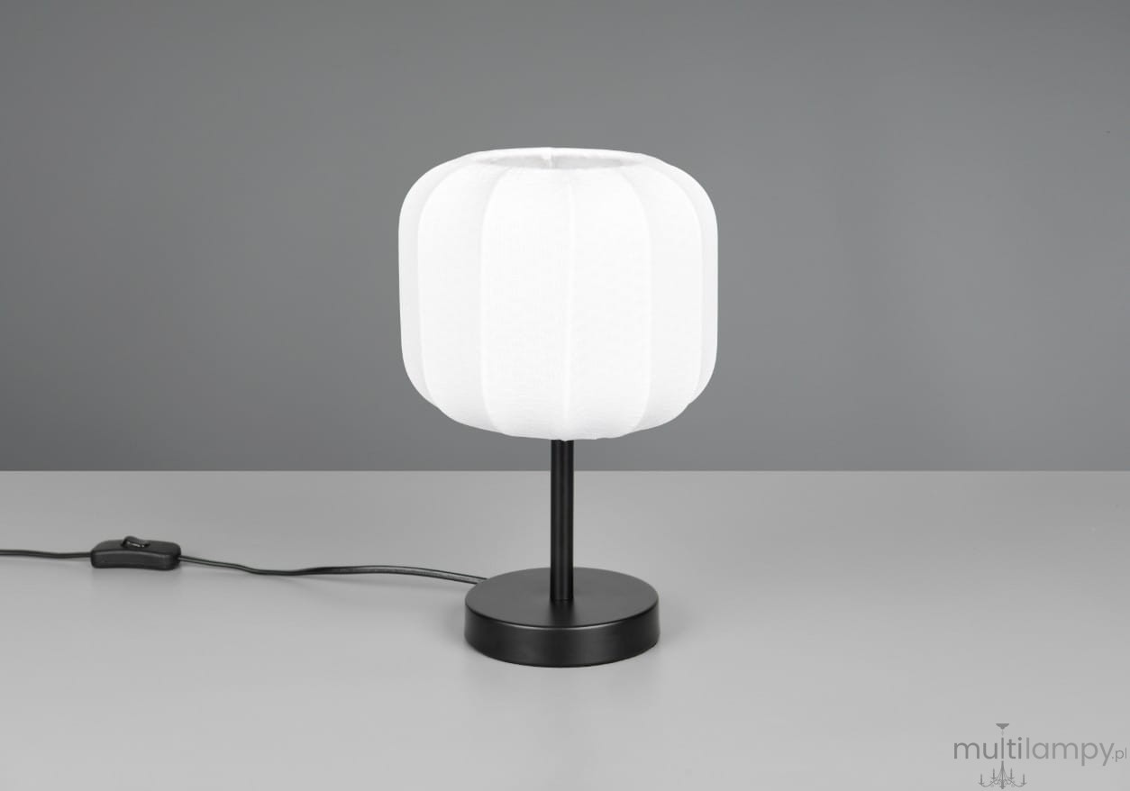 Lampa stołowa Madeline z białym kloszem w kształcie walca na czarnej podstawie. Ujęcie z boku, widoczny kabel z włącznikiem. Klosz ma delikatną fakturę, a całość ma nowoczesny design, idealna do wnętrz.