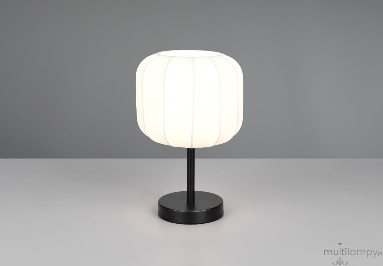 Lampa stołowa Madeline z białym kloszem w kształcie walca i czarną podstawą. Ujęcie frontalne, lampa stoi na szarej powierzchni. Widoczne delikatne linie na kloszu oraz prosty, minimalistyczny design.