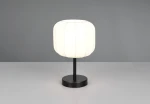 Lampa stołowa Madeline z białym kloszem w kształcie walca i czarną podstawą. Ujęcie frontalne, lampa stoi na szarej powierzchni. Widoczne delikatne linie na kloszu oraz prosty, minimalistyczny design.