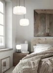 Lampa stołowa Madeline z czarną podstawą i białym kloszem, umieszczona na stoliku nocnym w sypialni. Klosz ma zaokrąglony kształt, a wokół widoczne są inne lampy wiszące oraz elementy drewniane w tle.