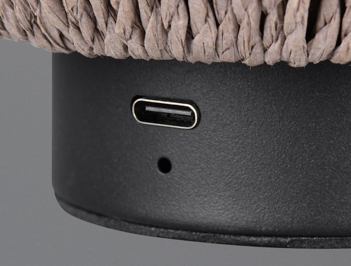 Zbliżenie na dolną część czarnej lampy stołowej Lira. Widoczny port USB-C do ładowania oraz detal brązowego materiału pokrywającego lampę. Tło szare, minimalistyczne.