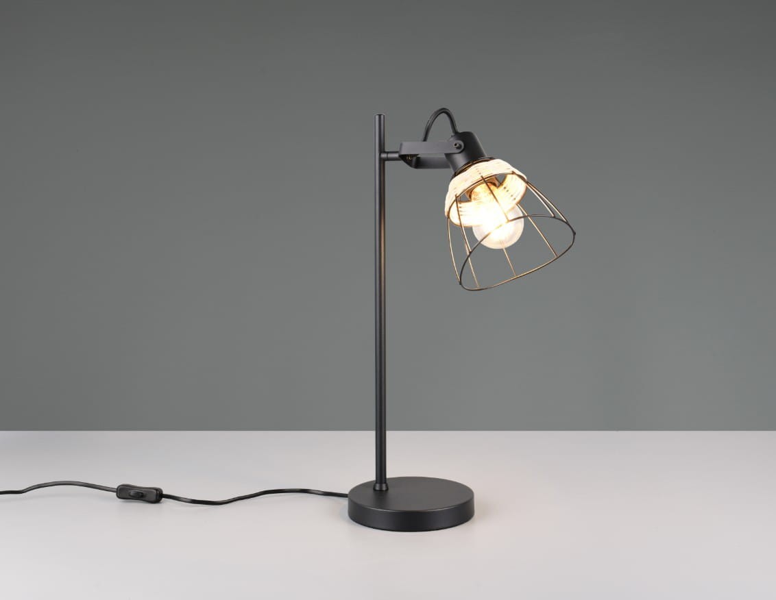 Lampa stołowa Jarome z czarnym metalowym stelażem i naturalnym kloszem w formie siatki. Ustawiona na białym blacie, z widocznym przewodem z włącznikiem. Klosz skierowany lekko w dół, eksponujący żarówkę E27. Tło w odcieniach szarości.