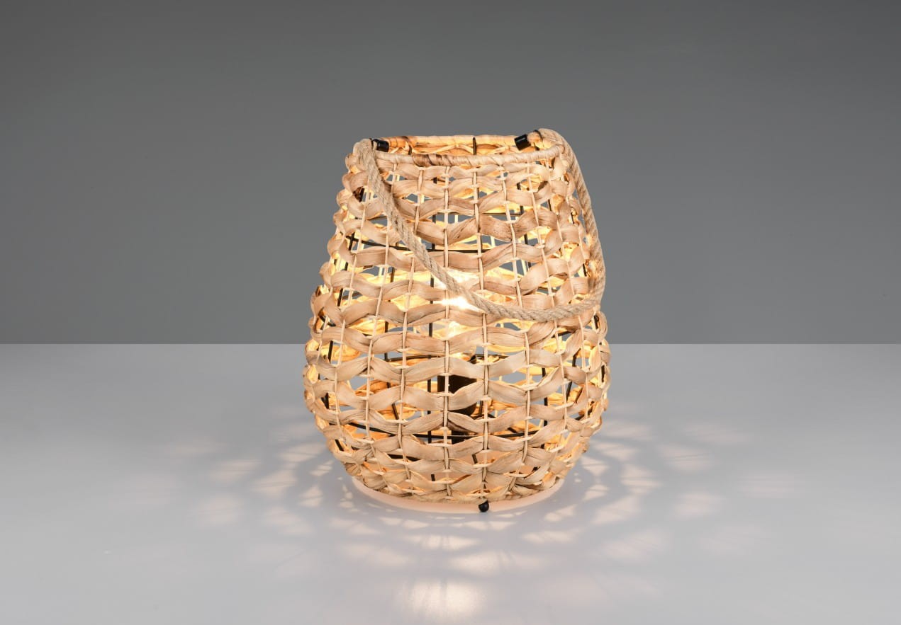 Lampa stołowa Jean w kolorze czarnym i naturalnym, wykonana z plecionki, o owalnym kształcie. Ujęcie frontalne, z widocznym źródłem światła wewnątrz. Lampa stoi na gładkiej, jasnej powierzchni, tworząc ciepłe światło i ciekawe wzory na tle.