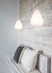 Lampa wisząca Janine w kolorze białym, w kształcie kropli, z teksturowanego materiału. Zawieszona na cienkim przewodzie, znajduje się nad łóżkiem z poduszkami na tle drewnianej ściany.