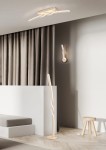 Kinkiet Ginko 1xLED/11W w piaskowym kolorze, zamontowany na ścianie, z unikalnym, skręconym kształtem. W tle zasłony w odcieniach szarości i minimalistyczne meble. Oświetlenie tworzy nowoczesną atmosferę.