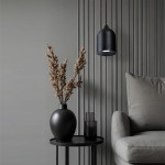 Czarna lampa wisząca Nilo z oprawą GU10, o zaokrąglonym kształcie, zawieszona nad stolikiem. W tle szare ściany z pionowymi panelami, obok wazon z suchymi gałązkami i szklane pojemniki. Mebel obity szaro-beżową tkaniną.