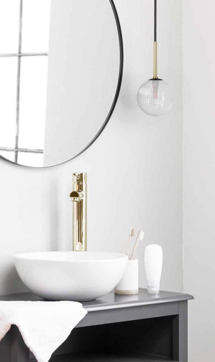 Lampa wisząca Aura 1xG9 w kolorze czarnym i transparentnym, z okrągłym kloszem w kształcie kuli. Zawieszona na cienkim przewodzie w złotym wykończeniu. W tle widoczna umywalka i lustro w okrągłej ramie.