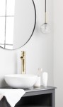 Lampa wisząca Aura 1xG9 w kolorze czarnym i transparentnym, z okrągłym kloszem w kształcie kuli. Zawieszona na cienkim przewodzie w złotym wykończeniu. W tle widoczna umywalka i lustro w okrągłej ramie.