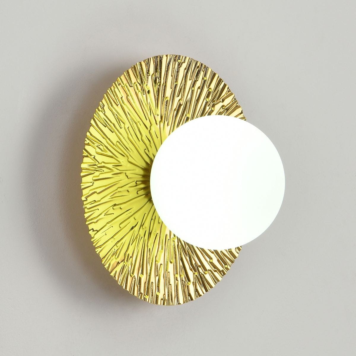 Kinkiet Sun S w kolorze złotym, o owalnej, teksturowanej podstawie z wypukłymi wzorami, z białą, okrągłą oprawą na żarówkę G9. Montowany na ścianie, widok frontalny na ścianie w jasnym otoczeniu.