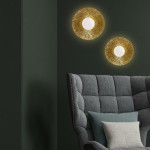 Kinkiet Sun S 1xG9 w kolorze złotym, okrągły z teksturowaną powierzchnią, z białym szklanym kloszem. Zawieszony na zielonej ścianie obok szarego fotela z poduszką, tworzy elegancki akcent w aranżacji wnętrza.