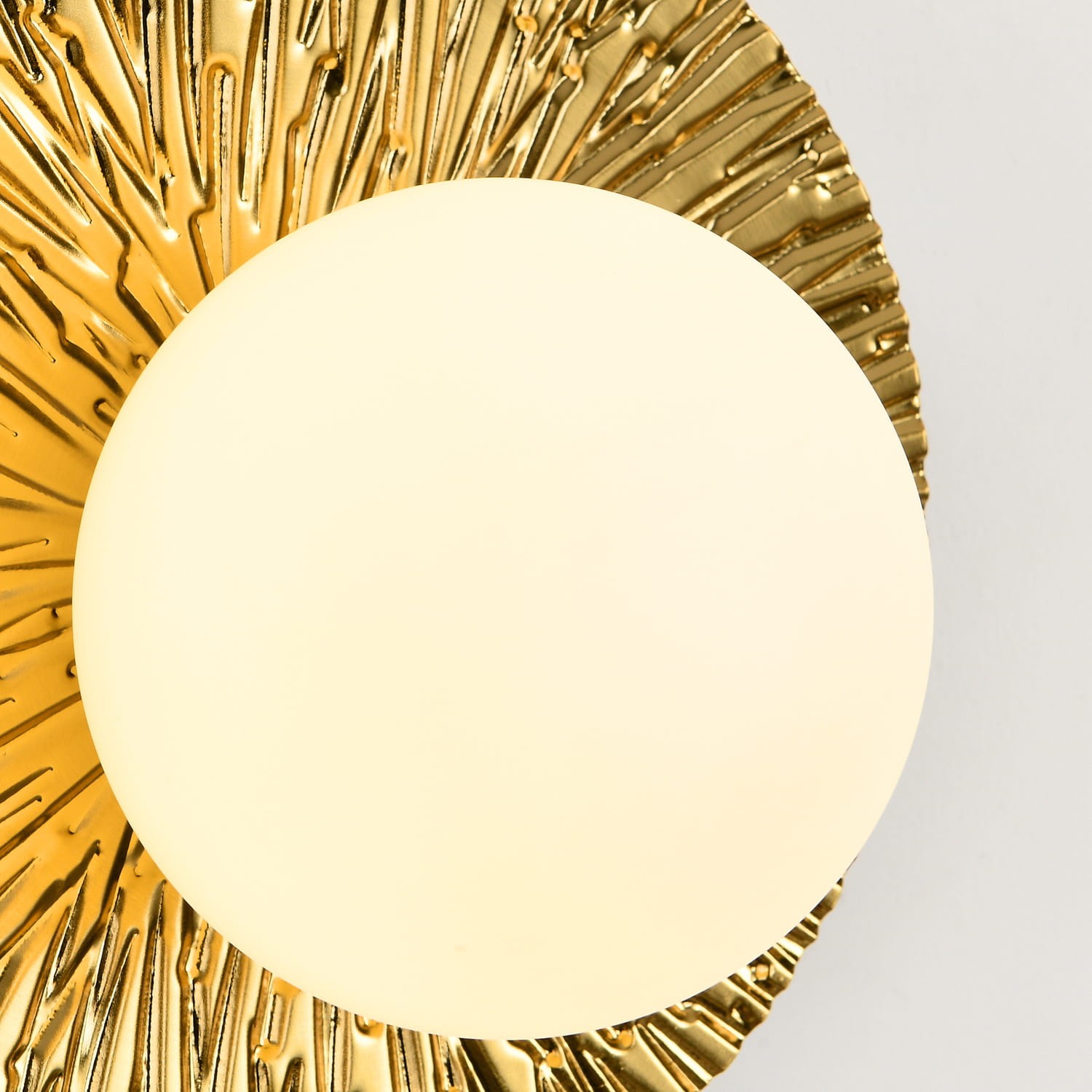 Kinkiet Sun M w złotym kolorze z teksturowaną, promienistą osłoną oraz okrągłym, matowym kloszem w białym kolorze. Produkt zamontowany na ścianie, widok z bliska, podkreślający detale wykończenia.