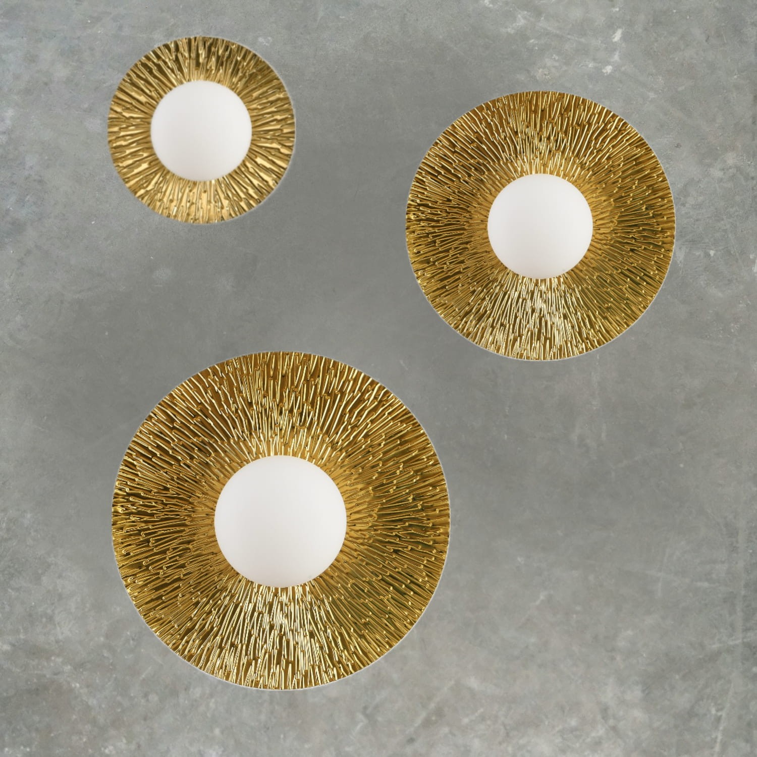 Kinkiet Sun L w kolorze złotym, z białym kloszem, o okrągłym kształcie. Zewnętrzna część ma teksturowaną, wypukłą strukturę. Widoczna jest grupa trzech lamp w różnych rozmiarach, zawieszonych na jednolitym tle.