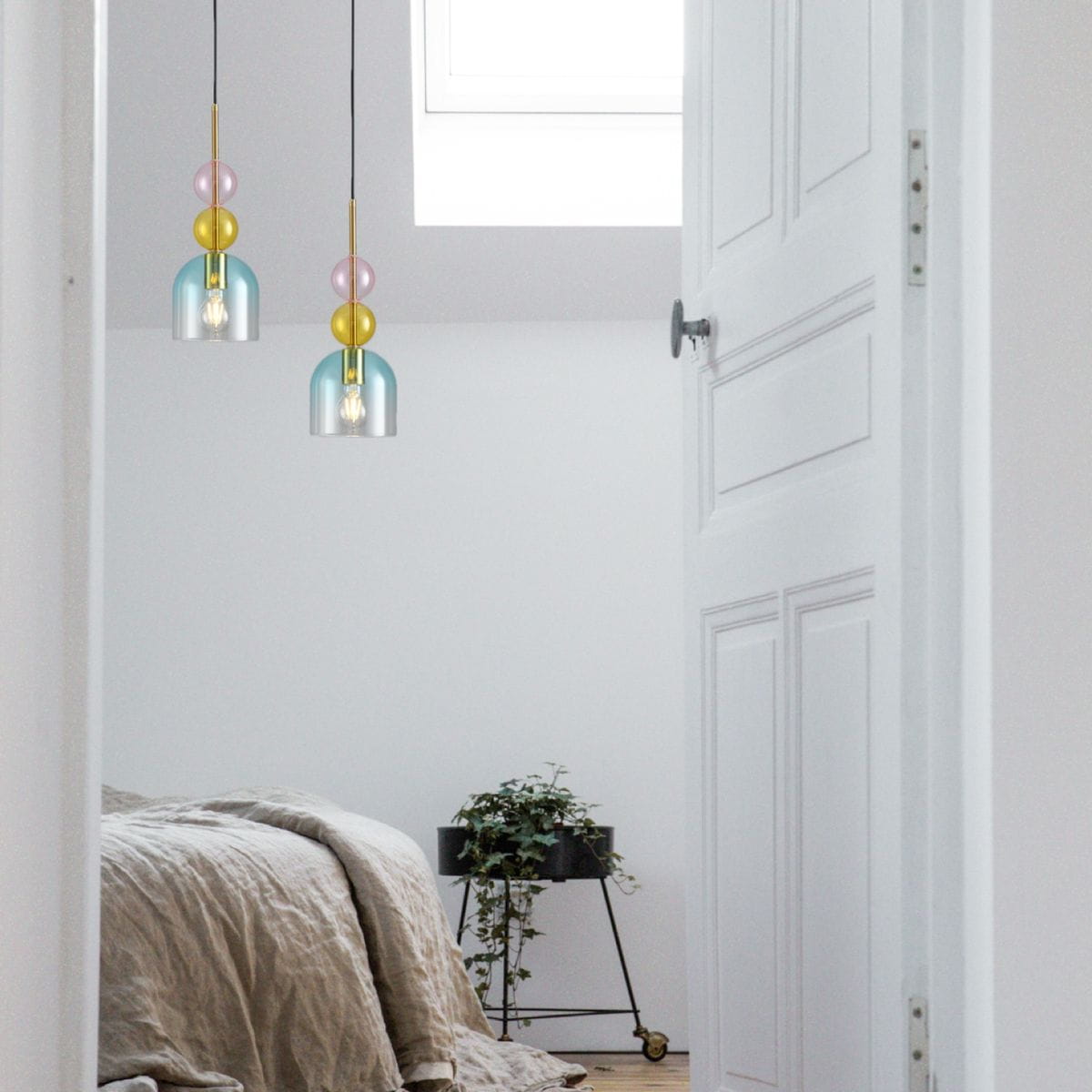Lampa wisząca Pastello 1xE27 w kolorze złotym i niebieskim, z przezroczystym kloszem oraz dekoracyjnymi kulami w odcieniach różu, żółci i niebieskiego. Zawieszona w jasnym pomieszczeniu, nad łóżkiem, obok rośliny w doniczce.