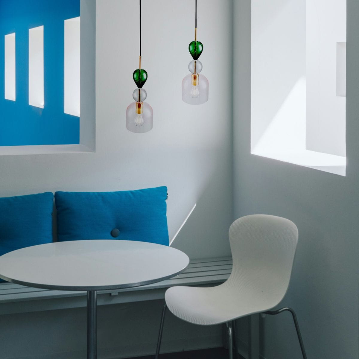 Lampa wisząca Pastello 1xE27 w kolorze złotym i różowym, z przezroczystymi kloszami. Zawieszona na czarnym przewodzie, w nowoczesnym wnętrzu z niebieskimi poduszkami. Klosze mają kształt dzwonka, eksponując źródło światła.