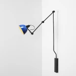 Kinkiet Aida z jedną oprawą E27 w kolorze czarnym i niebieskim. Lampa ma regulowany ramie, z zaokrągloną, niebieską czaszą o żółtym wnętrzu. Montaż na ścianie, na tle białej ściany.