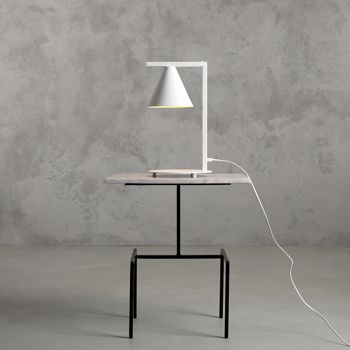 Lampa stołowa Form White z białym kloszem w kształcie stożka, umieszczona na prostokątnej, szarej podstawie z metalu. Stojak lampy ma minimalistyczny, prostokątny kształt. Widoczne przewody zasilające na stole z czarnymi nogami.