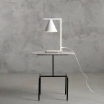 Lampa stołowa Form White z białym kloszem w kształcie stożka, umieszczona na prostokątnej, szarej podstawie z metalu. Stojak lampy ma minimalistyczny, prostokątny kształt. Widoczne przewody zasilające na stole z czarnymi nogami.