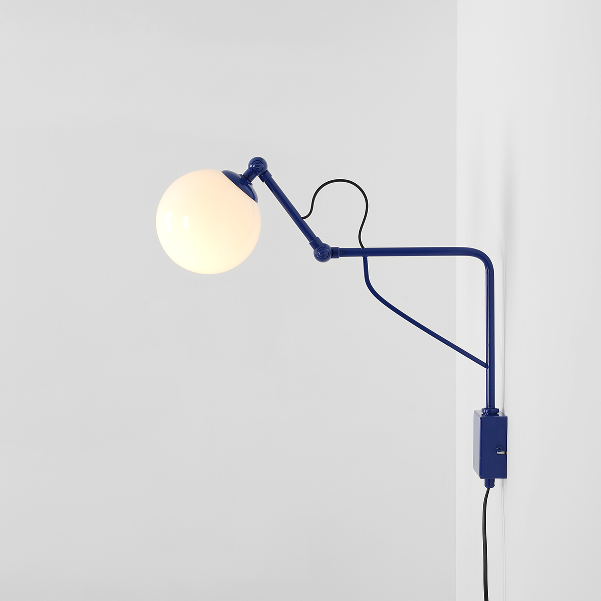 Kinkiet Soho Glass w kolorze kobaltowym z jedną oprawą E14. Lampa ma okrągłą, białą szklaną kulę oraz regulowany metalowy ramion, zamontowana na ścianie. Widoczny jest także czarny przewód i włącznik. Tło jasne.