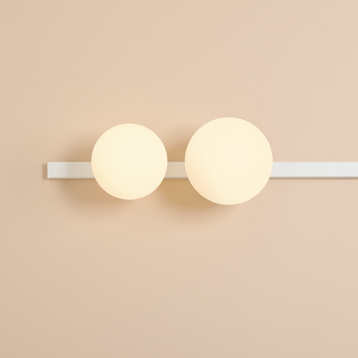 Biały kinkiet Garda z dwoma okrągłymi kloszami, jeden na żarówkę E14, drugi na E27, zamontowany na prostej, białej listwie. Klosze emitują miękkie światło, umieszczone na jasnym tle ściany.