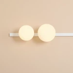 Biały kinkiet Garda z dwoma okrągłymi kloszami, jeden na żarówkę E14, drugi na E27, zamontowany na prostej, białej listwie. Klosze emitują miękkie światło, umieszczone na jasnym tle ściany.