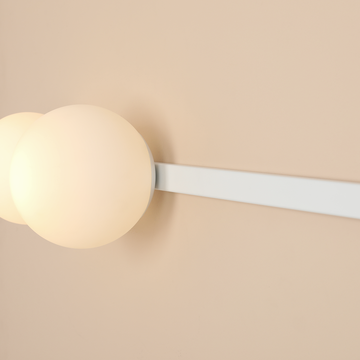 Biały kinkiet Garda z dwoma okrągłymi kloszami w kolorze mlecznym, zamontowany na prostokątnej białej podstawie. Lampa przeznaczona na żarówki E14 i E27, umieszczona na jasnobeżowej ścianie.