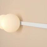 Biały kinkiet Garda z dwoma okrągłymi kloszami w kolorze mlecznym, zamontowany na prostokątnej białej podstawie. Lampa przeznaczona na żarówki E14 i E27, umieszczona na jasnobeżowej ścianie.