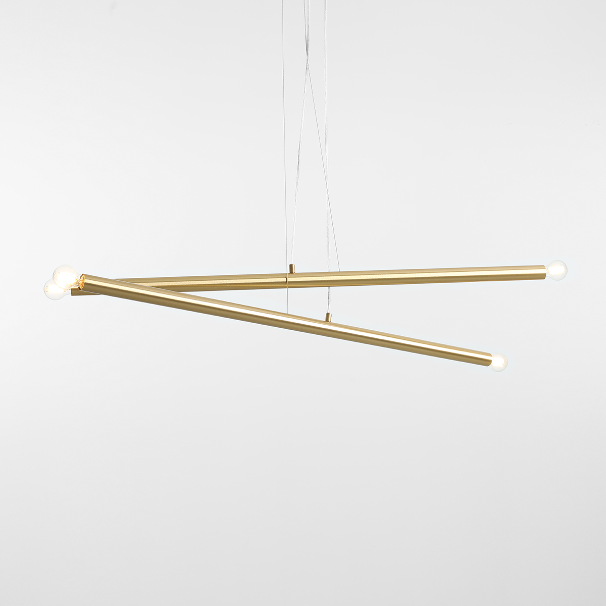 Lampa wisząca Tubo 4-punktowa w kolorze mosiądzu, składająca się z dwóch ukośnych, metalowych rur o matowej powierzchni. Z jednej strony umieszczone są dwa źródła światła, a z drugiej dwa gładkie zakończenia. Zawieszona na cienkich linkach na białym tle.
