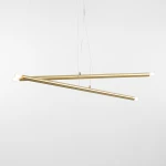 Lampa wisząca Tubo 4-punktowa w kolorze mosiądzu, składająca się z dwóch ukośnych, metalowych rur o matowej powierzchni. Z jednej strony umieszczone są dwa źródła światła, a z drugiej dwa gładkie zakończenia. Zawieszona na cienkich linkach na białym tle.