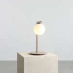 Beżowa lampa stołowa Zac z okrągłym kloszem, stojąca na okrągłej podstawie. Wysoka na około 40 cm, z metalowym stelażem i matową powierzchnią. Klosz emitujący ciepłe światło. Produkt na jednolitym tle, bez dodatkowych dekoracji.