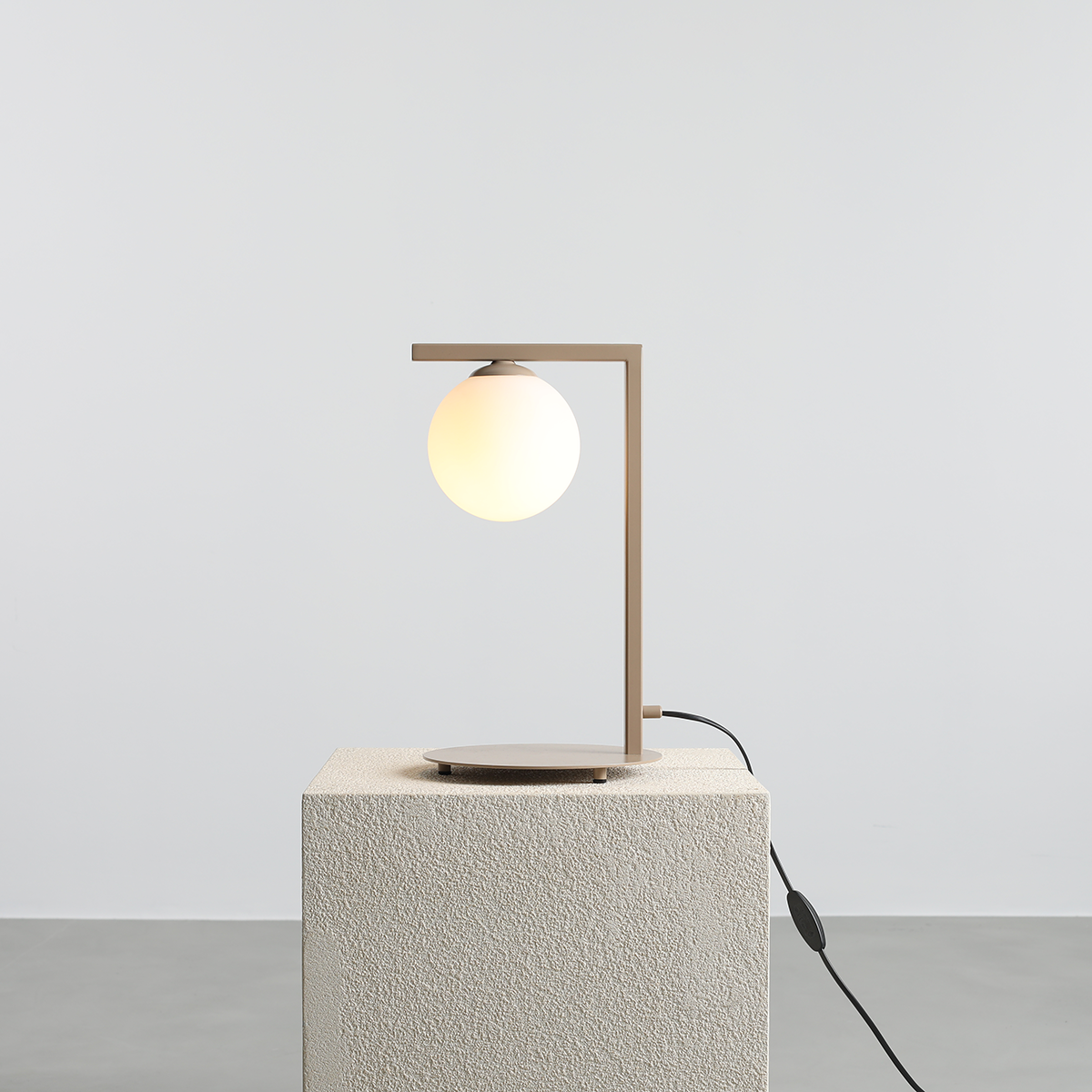 Beżowa lampa stołowa Zac z kulistym kloszem, umieszczona na okrągłej podstawie. Kąt ujęcia z boku, pokazujący prostą, nowoczesną formę oraz przewód z wyłącznikiem. Tło jasne, jednolite, podkreślające minimalistyczny design.