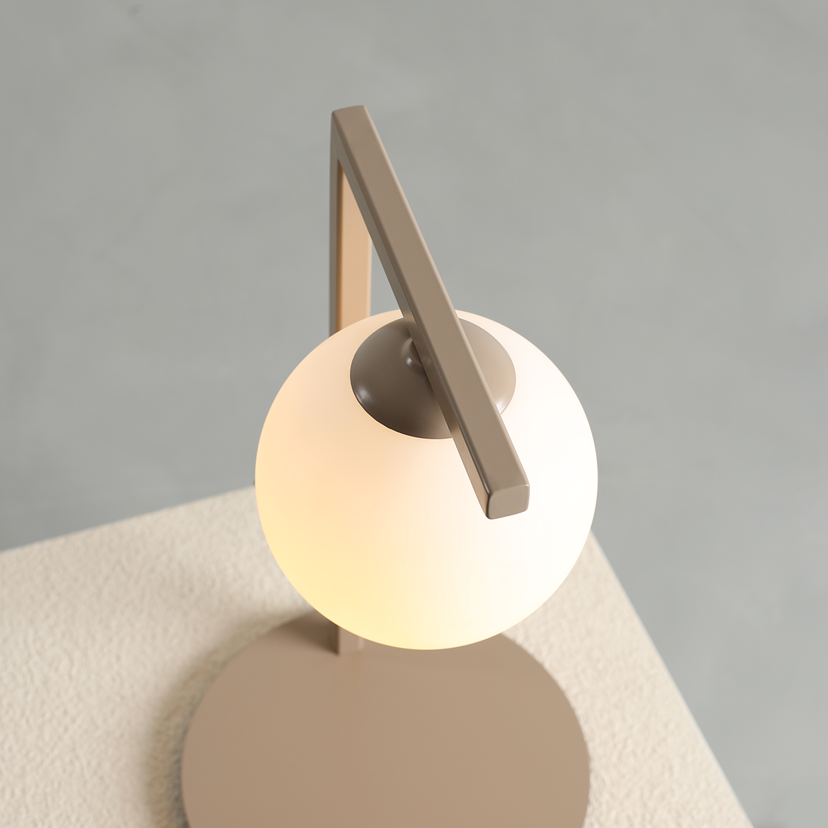 Beżowa lampa stołowa Zac z okrągłą, mleczną szklaną kulą i metalowym ramieniem w kształcie prostokąta. Lampa ustawiona na jasnej, teksturowanej powierzchni, widok z góry.