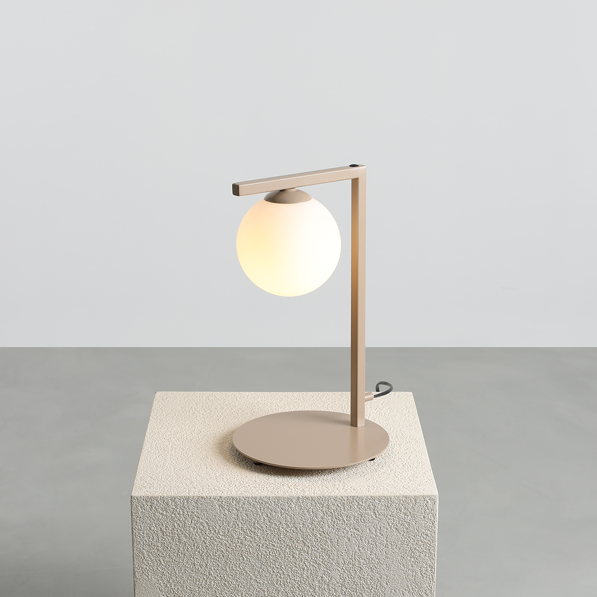 Beżowa lampa stołowa Zac z oprawą w formie prostokątnego ramienia i okrągłą, mleczną kulą. Ustawiona na kwadratowej, teksturowanej podstawie. Kąt ujęcia: frontalny, z wyraźnym ukazaniem detali.
