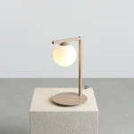 Beżowa lampa stołowa Zac z oprawą w formie prostokątnego ramienia i okrągłą, mleczną kulą. Ustawiona na kwadratowej, teksturowanej podstawie. Kąt ujęcia: frontalny, z wyraźnym ukazaniem detali.